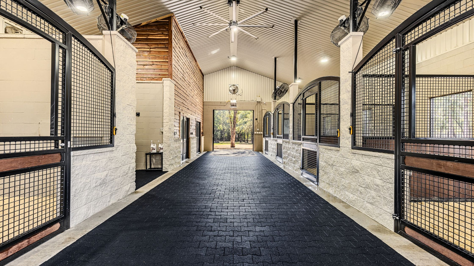 Elegant custom Horse Stalls. Long corridor with horse stables on either side in a well-lit indoor setting. Black Rubber pavers on aisle.  Arched "High" Euro Horse stall. Rectangular Yoke Opening with welded Mesh Door and Bottom shavings guard. Stall Fans and lighting. Jackson Ranch, TX.