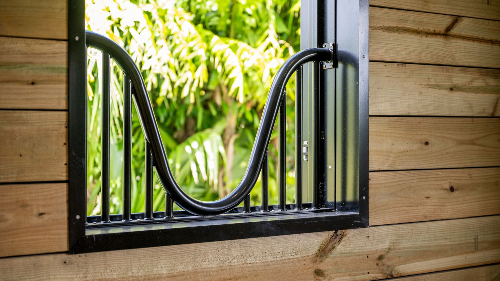 Custom-built Horse stable window. Solid welded Yoke Guard Window. 8M - Window Accessory.  Attached to a wooden wall with greenery in the background.